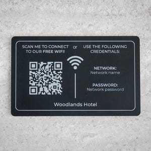 Op de afbeelding: Zwarte rechthoekige kaart met witte tekst en een QR-code. De kaart leest "SCAN ME OM VERBINDING TE MAKEN MET ONZE GRATIS WIFI!" en geeft netwerkgegevens. De hotelnaam "Woodlands Hotel" staat onderaan.