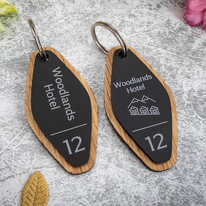 Pode incluir: Dois chaveiros de hotel de madeira com centro preto. Os chaveiros têm as palavras "Woodlands Hotel" e o número "12" impressos. Um chaveiro tem um gráfico de montanha e casa. Estão em um anel de metal prateado.