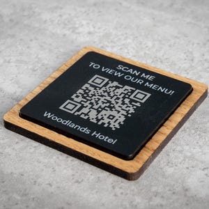 Può includere: Sottobicchiere quadrato in legno con una parte superiore nera con un codice QR e il testo "SCANSIONA PER VEDERE IL NOSTRO MENU!" e "Woodlands Hotel" in bianco. Il sottobicchiere è in legno marrone chiaro.