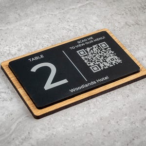 Puede incluir: Un letrero rectangular con el número de mesa con una superficie negra y un marco de madera. El letrero muestra el número "2" en plata, junto con un código QR y el texto "¡ESCANÉAME PARA VER NUESTRO MENÚ!" y "Woodlands Hotel".