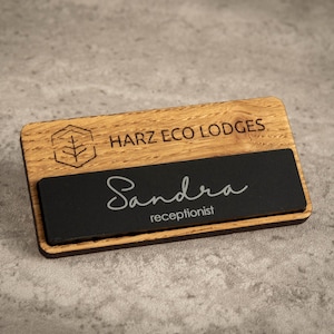 Könnte beinhalten: Namensschild aus Holz mit dem Text "HARZ ECO LODGES" und einem Logo. Der Name "Sandra" und die Berufsbezeichnung "Rezeptionist" sind auf einer schwarzen Platte dargestellt. Das Schild ist rechteckig und aus Holz.