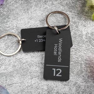 Pode incluir: Chaveiros pretos com argolas prateadas. Um chaveiro tem o texto "Recepção +1 234" e o outro diz "Woodlands Hotel" com o número "12". Os chaveiros estão sobre uma superfície cinza texturizada.