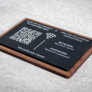 Könnte beinhalten: Eine rechteckige schwarze Karte mit Holzrahmen, die WLAN-Verbindungsdetails anzeigt. Die Karte zeigt einen QR-Code, ein WLAN-Symbol und den Text "Woodlands Hotel". Sie enthält Netzwerknamen und Passwortinformationen.