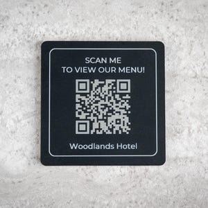 Puede incluir: Un posavasos cuadrado negro con texto blanco y un código QR. El texto dice "¡ESCANEAME PARA VER NUESTRO MENÚ!" y "Woodlands Hotel". El posavasos está sobre una superficie texturizada gris claro.