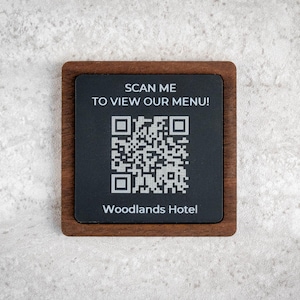 Può includere: Un cartello quadrato con un codice QR, sfondo scuro e cornice in legno. Il testo recita "SCAN ME TO VIEW OUR MENU!" e "Woodlands Hotel". Il cartello è su una superficie chiara e strutturata.
