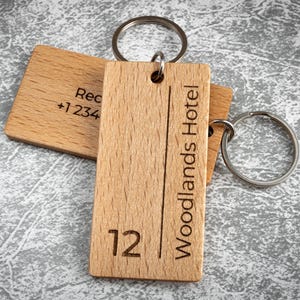 Pode incluir: Porta-chaves de madeira com argolas de metal. Um porta-chaves tem o texto "Woodlands Hotel" e o número "12". Outro porta-chaves tem o texto "Rec +1 234". Os porta-chaves são feitos de madeira clara e estão numa superfície cinzenta.