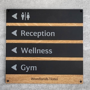 Könnte beinhalten: Rechteckiges Schild mit schwarzem Hintergrund und hellen Holzakzenten. Das Schild hat Richtungspfeile und Text für 'Rezeption', 'Wellness' und 'Gym'. Der obere Bereich zeigt ein Toilettensymbol. Am unteren Rand steht 'Woodlands Hotel'.