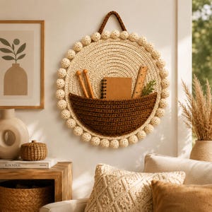 Puede incluir: Un organizador de pared circular hecho de cuerda trenzada color crema con un bolsillo de ganchillo marrón oscuro. El perímetro está decorado con pompones crema. El bolsillo contiene dos agujas de tejer de madera, una libreta y una regla.