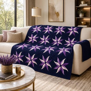 Könnte beinhalten: Eine dunkel marineblaue Steppdecke mit einem sich wiederholenden Muster aus achtstrahligen Sternmotiven in Violett-, Lavendel- und Weißtönen. Die Decke liegt über einem hellbeigen Sofa und zeigt aufwendige Steppnähte sowie ein geometrisches Blumenmuster.