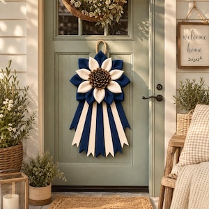 May include: A decorative door hanging featuring a large, layered flower design with navy blue, cream, and brown elements. The flower is attached to a navy blue ribbon with cream stripes. A welcome sign hangs nearby.