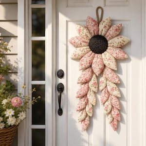Pode incluir: Um enfeite de parede em forma de girassol feito de tecido, com um centro texturizado castanho-escuro e inúmeras pétalas almofadadas em tecido creme com um padrão floral rosa. A flor tem uma longa cauda de pétalas e uma alça de juta.