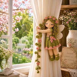 May include: A crochet fairy doll curtain tieback featuring a figure with wavy green hair, a green skirt, and pink flower accents. The doll has white wings and holds a long, trailing vine of green leaves and pink flowers that wraps around a white curtain.