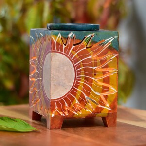 Op de afbeelding: Een vierkante stenen wierookbrander met een gegraveerd zonontwerp. De brander is geschilderd in tinten rood, oranje, geel en groen. Hij heeft een klein, rond putje in het midden om wierook te branden.