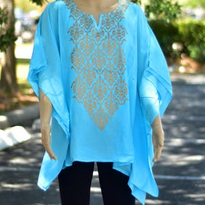 May include: A light blue tunic top with a gold-colored, ornate pattern on the front. The tunic has a V-neck and wide, flowing sleeves. The garment is made of a lightweight fabric.