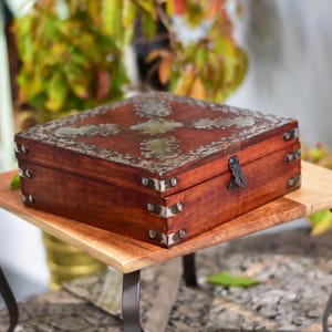 May include: A square wooden box with a dark reddish-brown finish, adorned with silver-colored metal accents. The box features a decorative metal design on the lid and metal corner protectors. A black latch secures the box, which sits on a light brown wooden surface.