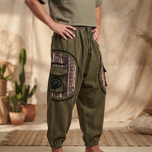 May include: Olive green trousers featuring an elastic waistband with a drawstring, cuffed ankles, and large side cargo pockets. The pockets are accented with vertical multicolored striped fabric panels and a black spiral embroidered design.