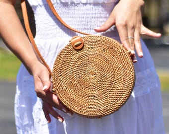 Op de afbeelding: Een ronde, geweven rotan tas met een bruine leren riem en sluiting. De tas wordt tegen een witte jurk gehouden, waarbij de natuurlijke, aardse tinten worden benadrukt. Het cirkelvormige ontwerp en de textuur van de tas zijn prominent aanwezig.