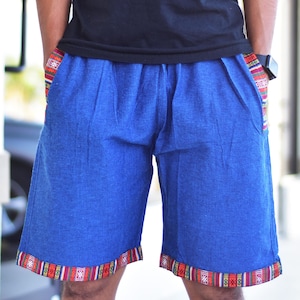 May include: Blue shorts with patterned trim on the pockets and hem. The shorts are made of a solid blue fabric and have a relaxed fit. The trim features a colourful geometric design with red, orange, and green accents. The shorts have pockets.