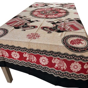 May include: Beige tablecloth with a repeating pattern of elephants, floral designs, and circular motifs in red and black. The tablecloth has a black border and is draped over a table.