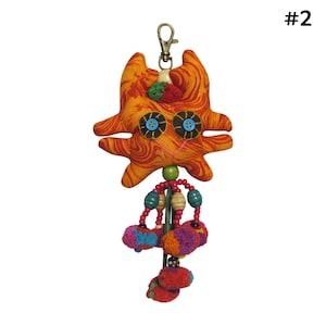 May include: Orange and red fabric cat keychain with blue eyes and a pink smile. The keychain has a wooden bead and colorful pom-poms hanging from it.