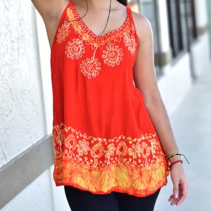 Könnte beinhalten: Ein ärmelloses Top mit V-Ausschnitt in leuchtendem Rot, verziert mit aufwendigen weißen und orangefarbenen Blumenmustern. Der untere Saum hat ein gelbes und orangefarbenes Batik-Design. Das Top ist aus leichtem Stoff gefertigt.