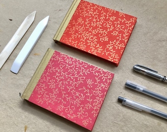 May include: Two small, square journals with gold-colored spines and covers with a floral pattern in gold. One journal is coral, the other is pink. Two bone folders and two pens are also visible.