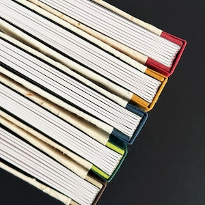 May include: A stack of journals with colorful spines. The journals have white lined pages and are bound with fabric in various colors, including red, yellow, blue, and green. The background is a solid black.