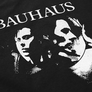 May include: Black t-shirt featuring a white graphic of two faces and the word "BAUHAUS" in bold, capital letters. The design has a vintage, distressed aesthetic. The faces are in a high-contrast, black and white style.