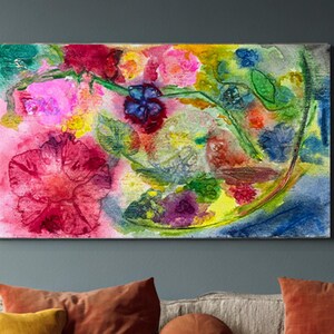 May include: A vibrant watercolor painting of flowers in shades of pink, red, yellow, and blue. The artwork hangs above a beige sofa with colorful throw pillows. A small wooden side table and a vase with branches complete the cozy living room scene.
