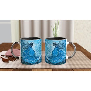 Könnte beinhalten: Zwei weiße Keramikbecher mit schwarzen Rändern. Beide Becher zeigen eine schwarze Katze mit blauen und weißen wirbelnden Designs. Die Katze ist mit einem fließenden blauen Gewand und einem weißen Hintergrund dargestellt.