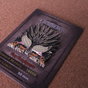 May include: A silver and black enamel pin featuring a monster with a spiky back and a red and white face. The pin is on a card with the text "Enter of Elders" and "Slay Nergigante". The card also includes the text "Reward Money: 18000z" and "Time Limit: 50 min."