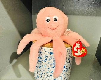 May include: Pink octopus beanie baby plush toy with a white tag that says "Ty" and a red heart with a star. The toy is sitting on a white and blue ceramic container.