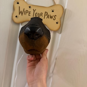 Puede incluir: Una escultura de pared con forma de nariz de perro, con trufa negra y hocico marrón, situada frente a un letrero de madera claro con forma de hueso que incluye el texto "WIPE YOUR PAWS" en letras negras y pequeños dibujos de huellas.