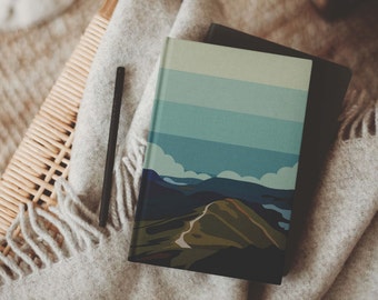 May include: A notebook with a landscape illustration on the cover. The illustration features a green hill with a winding road, blue mountains in the background, and a light blue sky with white clouds.