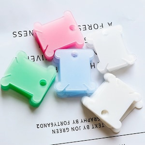 May include: A set of six thread bobbins in various colours, including pink, green, light blue, and white. The bobbins are stacked on top of each other and are made of clear plastic.