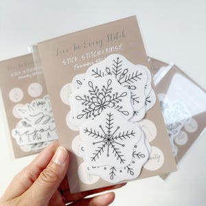 May include: A set of white felt snowflake embroidery patterns with black outlines. The patterns are packaged in a clear plastic sleeve with a brown card backing. The card has the text "Love In Every Stitch" and "Stick Stitch & Rinse" printed on it.