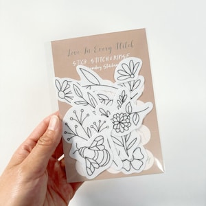 Pode incluir: Um conjunto de adesivos brancos para aro de bordado com desenhos de linha preta de flores e uma abelha. Os adesivos dizem "Love in Every Stitch" e "Stick Stitch & Rings Embroidery Stabilizer".