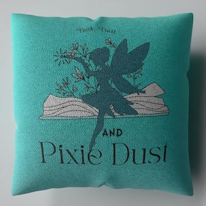 Puede incluir: Un cojín cuadrado de color verde azulado con la silueta oscura de un hada alada sobre un libro abierto. El texto "Faith, Trust" aparece sobre el hada, y "AND Pixie Dust" está impreso en letras negras debajo de la ilustración del libro.