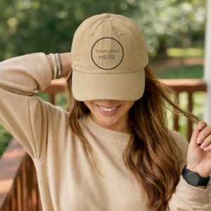 May include: A tan baseball cap featuring a centered black circular graphic with the text YOUR LOGO HERE. The cap has a structured crown, a curved brim, and a fabric strap with a metal buckle closure at the back for an adjustable fit.