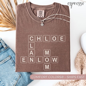 May include: A brown Comfort Colors t-shirt with the name "Chloe" spelled out in Scrabble tiles. The shirt also has the name "M" and "Enlow" spelled out in Scrabble tiles. A silver necklace with a small pendant is draped across the shirt.