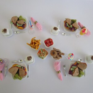 May include: A collection of miniature food items including sandwiches on square plates, bowls of fries, pasta, and pretzels, accompanied by small white cups, silver-toned cutlery, and pink napkins with gold rings, all arranged on a plain white surface.