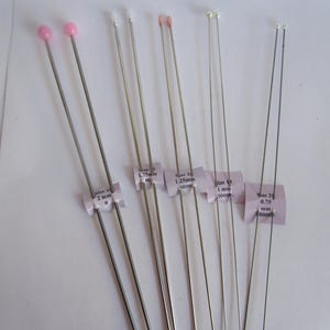 May include: A collection of sewing pins with various head colors, including pink, white, and pearl. The pins are of different sizes, with labels indicating measurements in millimeters. The pins are arranged on a white surface.
