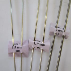 May include: Close-up of several silver knitting needles with pink paper tags indicating sizes: 1.5 mm, 1.75 mm, and 2 mm. The needles are thin and metallic, ideal for fine yarn projects.