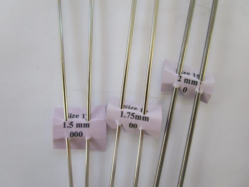 May include: Close-up of several silver knitting needles with pink paper tags indicating sizes: 1.5 mm, 1.75 mm, and 2 mm. The needles are thin and metallic, ideal for fine yarn projects.