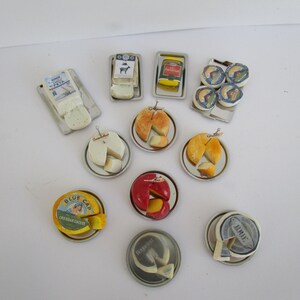 Può includere: Una collezione di repliche di formaggio in miniatura disposte su piccoli vassoi e piatti di metallo. Il set include vari tipi di formaggio come feta, formaggio erborinato, brie e forme rivestite di cera, alcune con etichette come Blue Cap e Brie.
