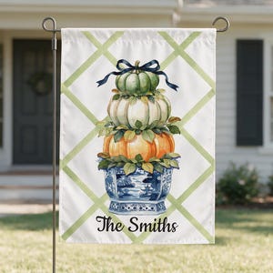 May include: A rectangular garden flag featuring a white background with a light green lattice pattern. The centre displays a stack of three pumpkins in green, white, and orange, topped with a black bow, sitting in a blue and white chinoiserie planter. The text "The Smiths" is printed in black script at the bottom.