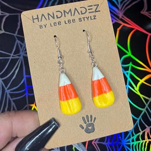 May include: A pair of dangle earrings with a candy corn design. The earrings are silver and have a white, orange, and yellow candy corn charm. The earrings are on a brown card with the text "HANDMADDEZ BY LEE LEE STYLZ".