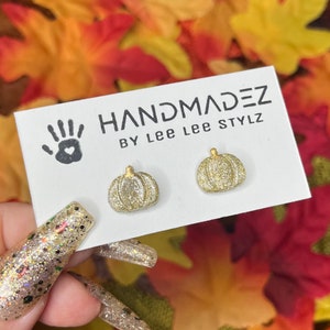 May include: A pair of gold glitter pumpkin earrings. The earrings are on a white card with the text "HANDMADEZ BY Lee Lee STYLZ". The background is a blurred image of fall leaves.