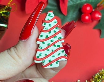 May include: A white Christmas tree charm with red and green stripes and green glitter. The charm is held by a hand with red nail polish.