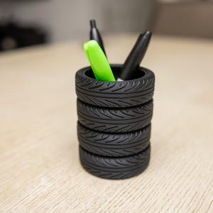 May include: A black tire pen holder with four stacked tires. The holder contains a green highlighter and two black pens.